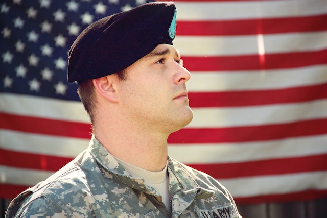 A military service member wearing fatigues and a black beret standing in front of an American flag